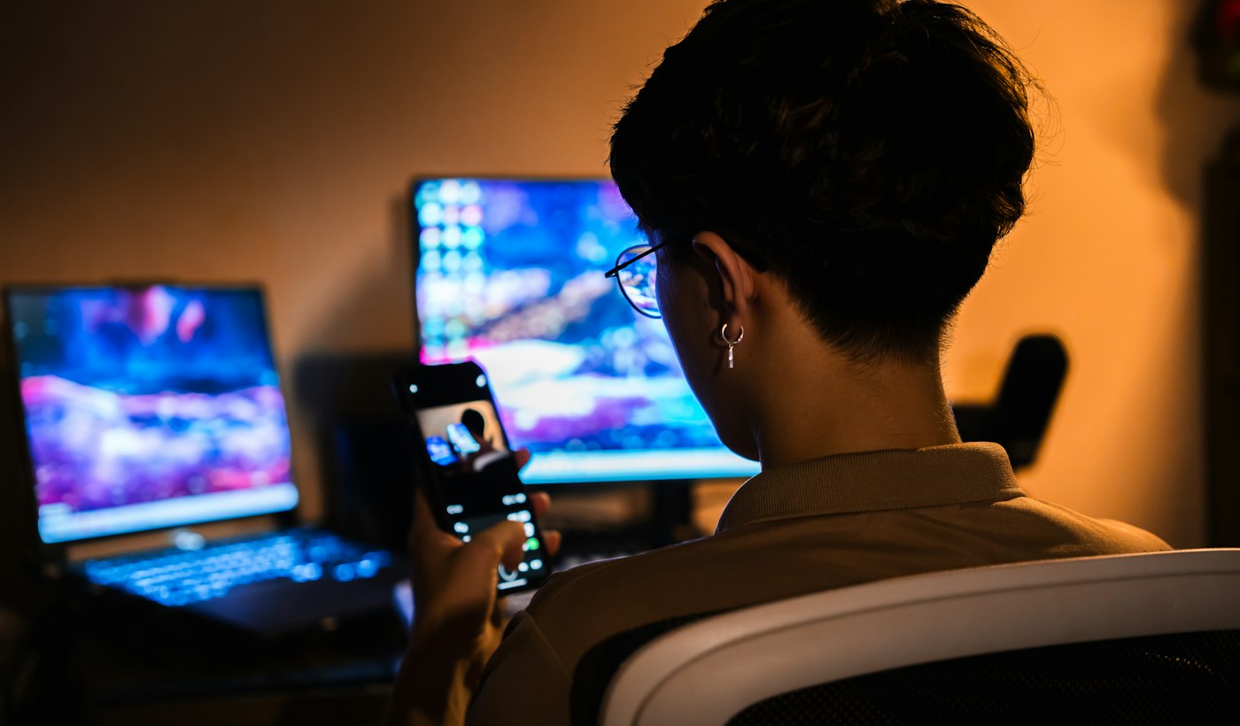 A person using a phone in front of glowing computer screens in a dark room