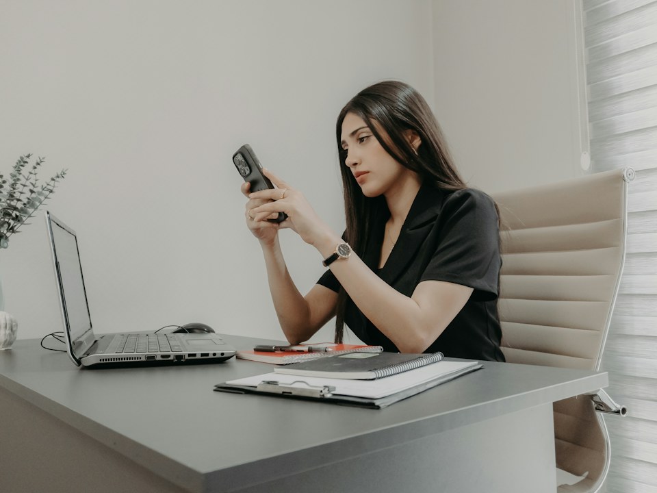 A woman looking at her phone at a desk with a laptop