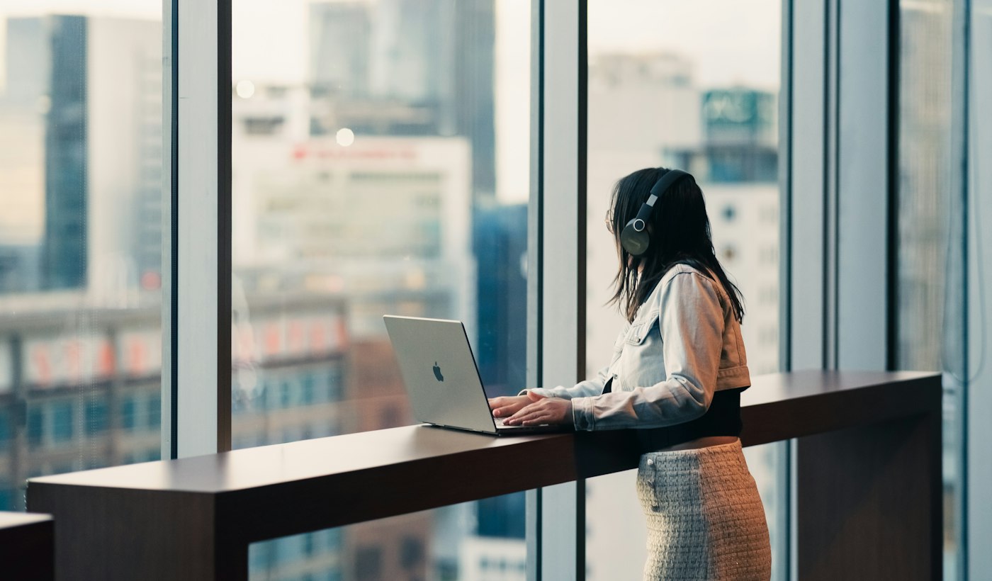 A person wearing headphones working on a laptop beside a window