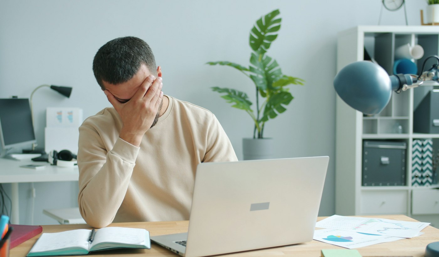 A person alone at a desk rubbing their face in front of a laptop